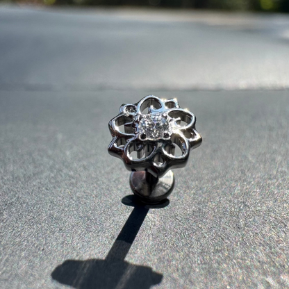 Silver flower-shaped earring on a reflective surface with blurred greenery in the background